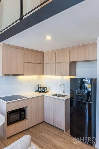 Condominium for rent in Khlong Toei, Bangkok - Modern light‑wood kitchen with induction hob, microwave and black fridge beneath a mezzanine.