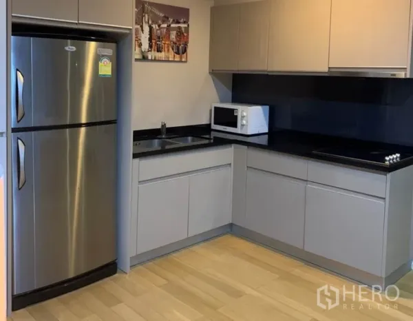 Condominium for rent in Watthana, Bangkok - Modern L-shaped kitchen with black countertops, stainless refrigerator, cooktop and microwave.