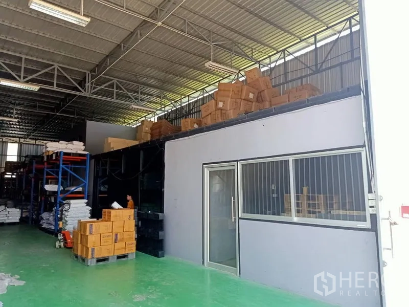 Warehouse for rent in Bang Kapi, Bangkok - Close view of the mezzanine office with windows inside the warehouse, with boxes stacked on racks under a metal roof.