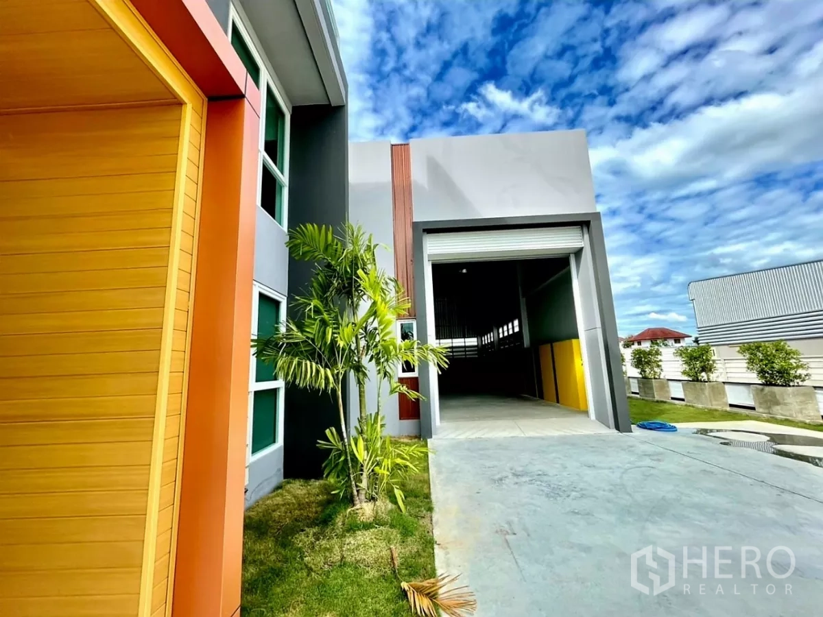 Warehouse for sale in Lam Luk Ka, Pathum Thani - Warehouse loading bay with roller shutter beside a landscaped office entrance and palm tree.