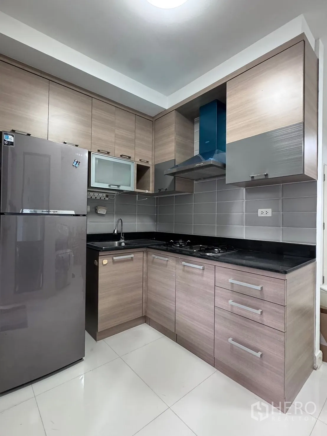 Townhouse for rent in Phra Khanong, Bangkok - L-shaped kitchen with gas hob, hood, fridge, and ample cabinets.