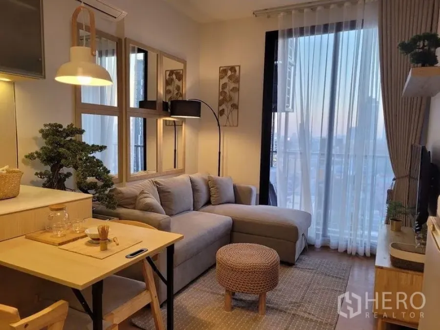 Condominium for sale in Watthana, Bangkok - Open-plan living area with dining nook, balcony doors, and warm wood finishes at dusk.