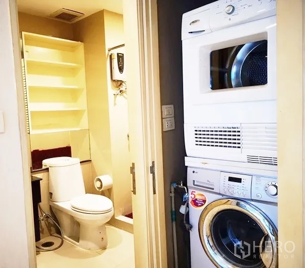 Condominium for rent in Khlong Toei, Bangkok - Laundry nook with stacked washer and dryer beside a guest bathroom with shelving.