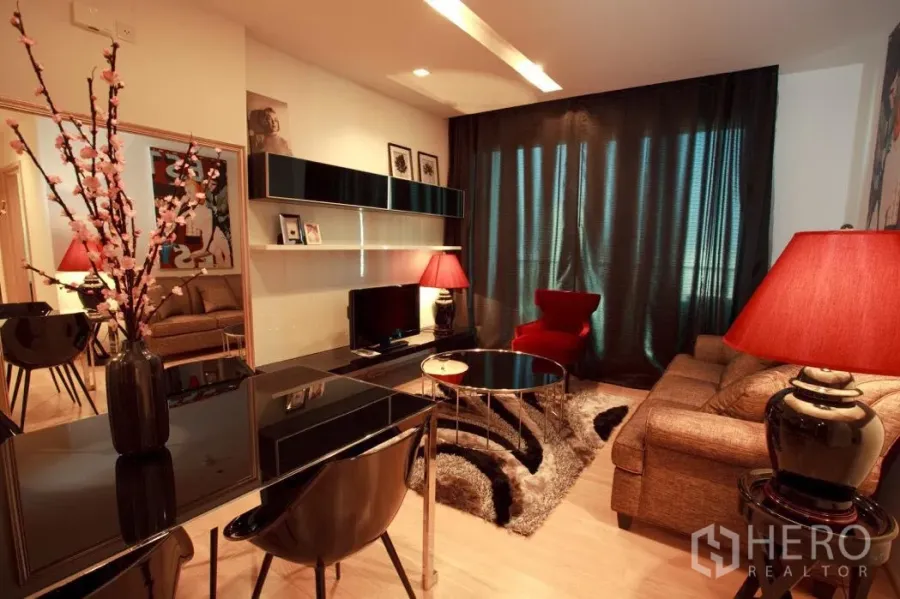 Condominium for rent in Khlong Toei, Bangkok - Warm living room with dark curtains, sectional sofa, red accent chair, TV console and floating shelves.