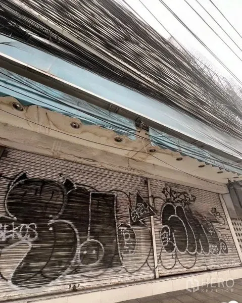 店屋 出租在Watthana, Bangkok - 更广角的店面外立面，蓝色雨棚下方为卷帘门与大量线路。