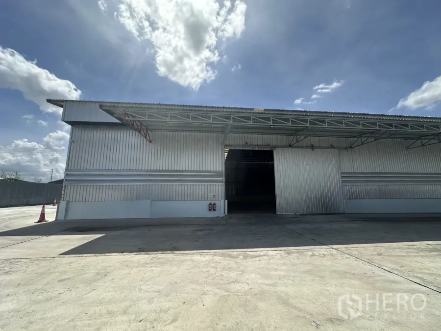 货栈 出租在Mueang Pathum Thani, Pathum Thani - 金属板外立面的仓库正面，宽幅滑动门、装卸雨棚与宽阔混凝土庭院，晴空之下。