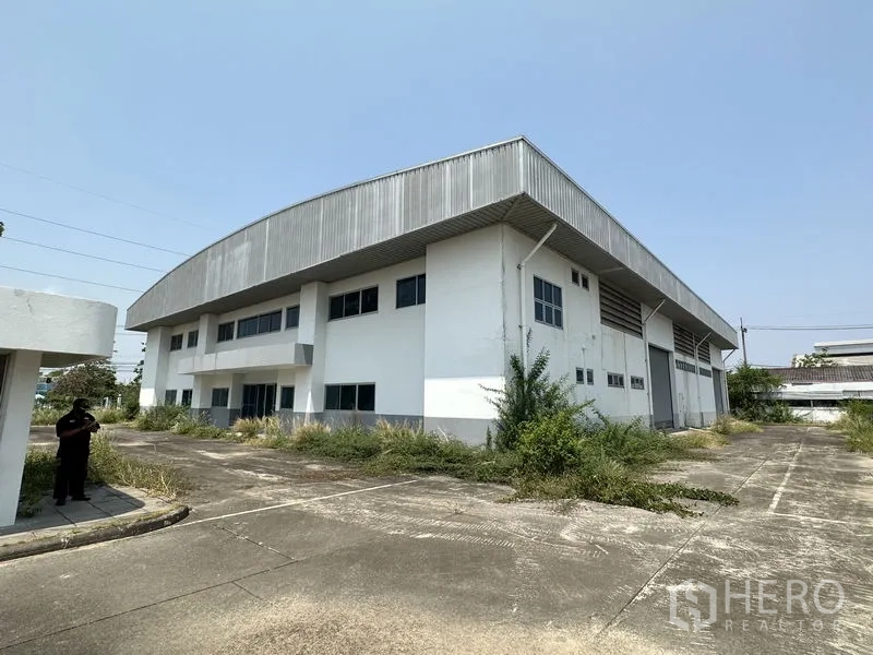 Factory for rent in Chon Buri - Angled exterior of the two‑storey factory showing wide concrete driveway and overhanging roof in a landscaped yard.