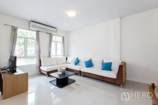 联排别墅 出租在Watthana, Bangkok - 采光充足的客厅，转角沙发、空调与大窗户，面向前院。