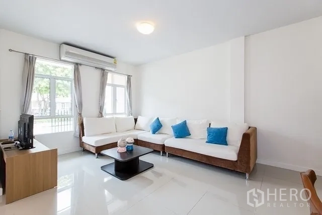 联排别墅 出租在Watthana, Bangkok - 采光充足的客厅，转角沙发、空调与大窗户，面向前院。