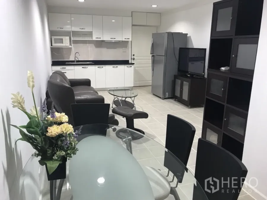 Condominium for rent in Khlong Toei, Bangkok - Open-plan living and dining area with glass table, leather sofa, TV unit, and white kitchenette with microwave and fridge.