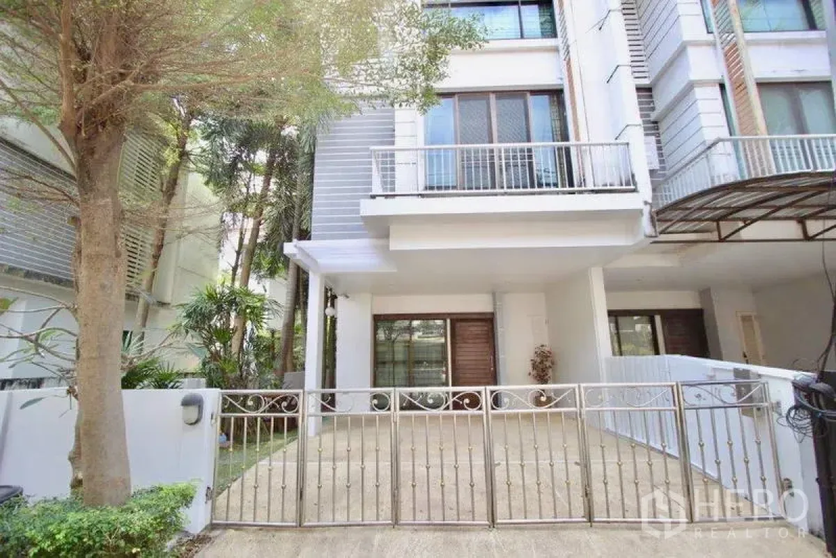 Townhouse for sale in Suan Luang, Bangkok - Modern three-storey townhouse facade with gated driveway and balcony, shaded by trees.