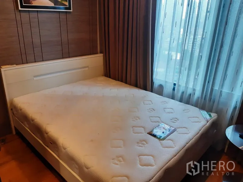 Condominium for rent in Khlong Toei, Bangkok - Cozy bedroom with large bed, textured accent wall and a full-height window with sheer curtains.