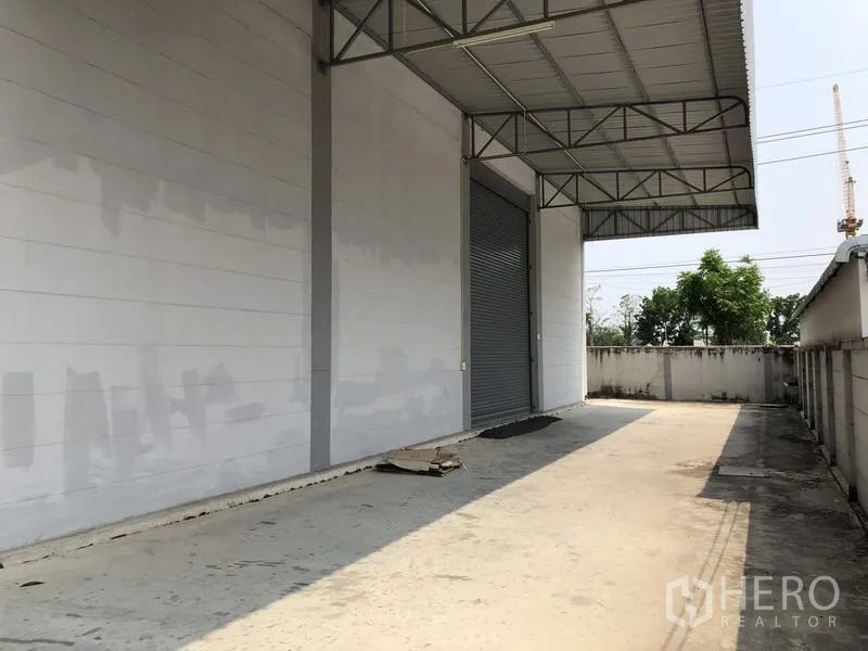 Factory for rent in Lam Luk Ka, Pathum Thani - Covered loading bay with metal canopy, roller shutter and wide concrete apron beside the perimeter wall.