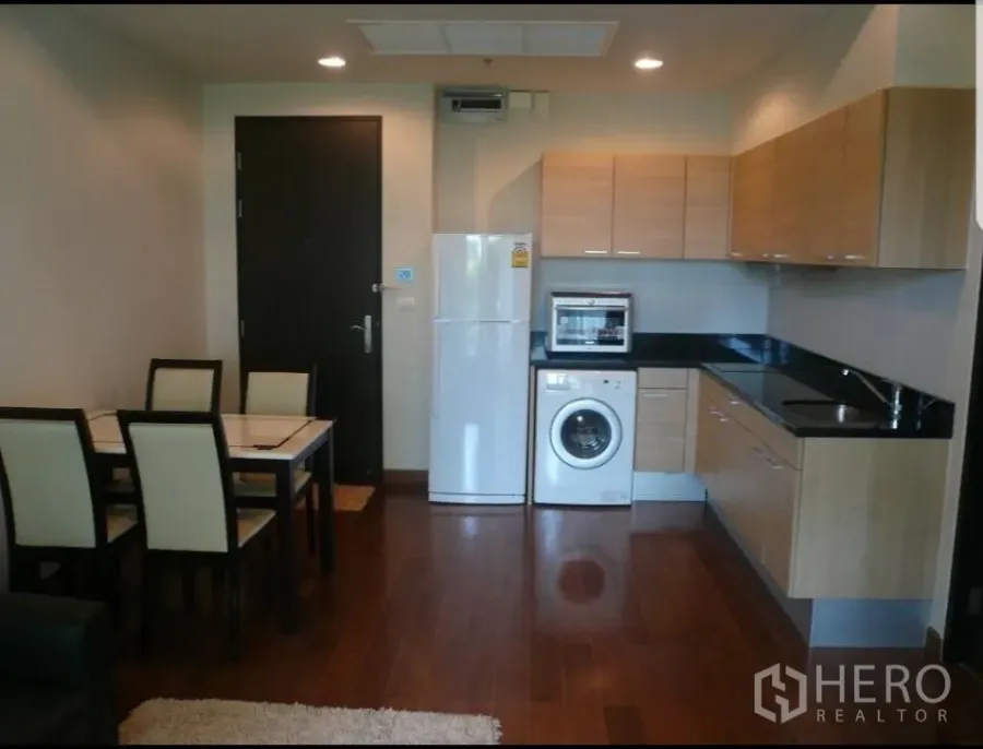 Condominium for rent in Pathum Wan, Bangkok - Open-plan kitchen with wood cabinets, black countertops, fridge, washing machine and dining set on hardwood floors.
