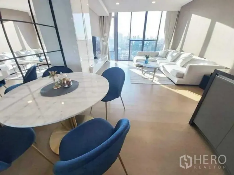 Condominium for rent in Khlong Toei, Bangkok - Dining area with round marble table and blue chairs beside sunny living room.