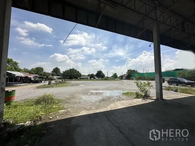 Factory for rent in Phra Nakhon Si Ayutthaya - View from under the canopy across an open gravel yard toward the site entrance and trees.
