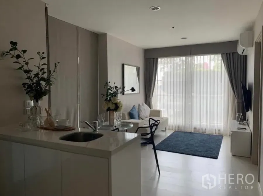 Condominium for rent in Khlong Toei, Bangkok - Open-plan living room with white kitchenette island, sofa, and floor-to-ceiling windows with sheer curtains.