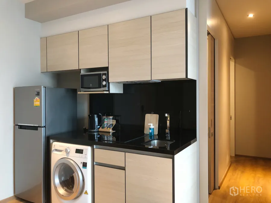 Condominium for rent in Khlong Toei, Bangkok - Modern kitchenette with light wood cabinets, black countertop, refrigerator and in-unit washing machine.