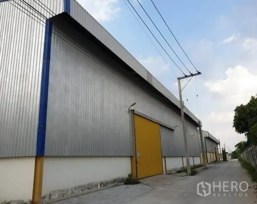 Warehouse for rent in Lat Krabang, Bangkok - Side exterior of corrugated metal warehouse with yellow roller doors along a quiet service road and power lines.