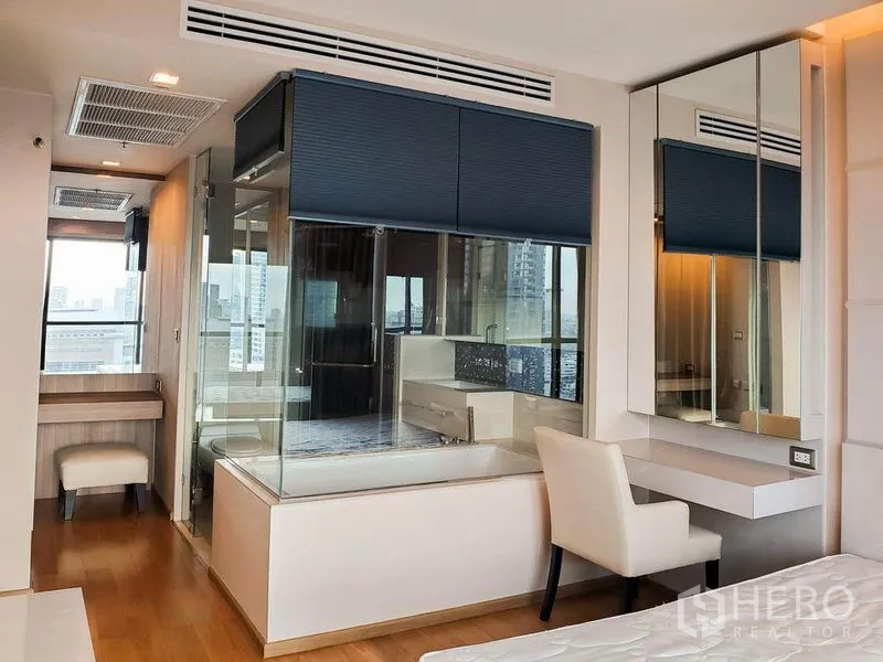 Condominium for rent in Bang Rak, Bangkok - Glass-walled ensuite bathroom with bathtub, large mirrors, and a vanity desk adjoining the bedroom.