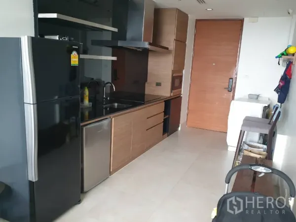 Condominium for rent in Khlong Toei, Bangkok - Modern kitchenette featuring a black refrigerator, built-in wooden cabinets, sink, and entry door.