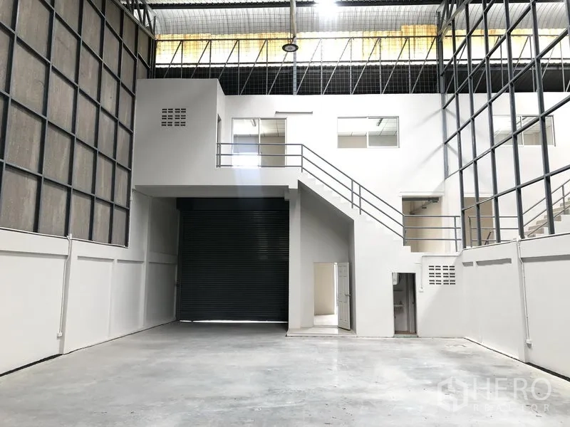 Factory for rent in Pak Kret, Nonthaburi - Warehouse interior showing mezzanine offices, staircase and a large roller shutter loading bay.