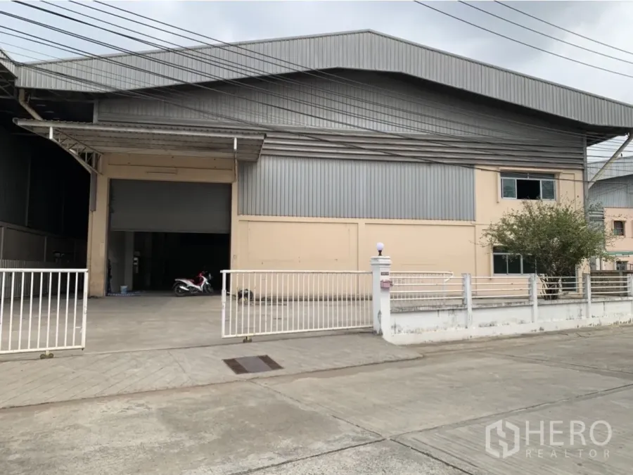 Factory for rent in Mueang Samut Prakan, Samut Prakan - Gated factory exterior with metal cladding, wide driveway and covered loading bay in an industrial area.