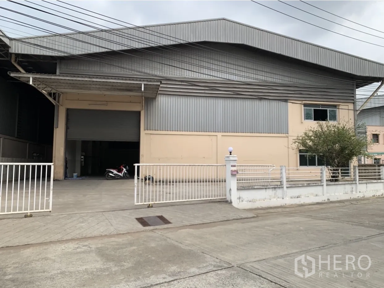 Factory for rent in Mueang Samut Prakan, Samut Prakan - Gated factory exterior with metal cladding, wide driveway and covered loading bay in an industrial area.