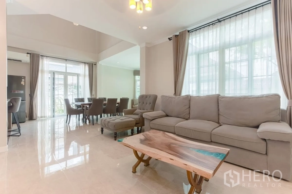 Detached House for rent in Bang Na, Bangkok - Living room with large windows, sheer drapes, and seating next to dining zone.