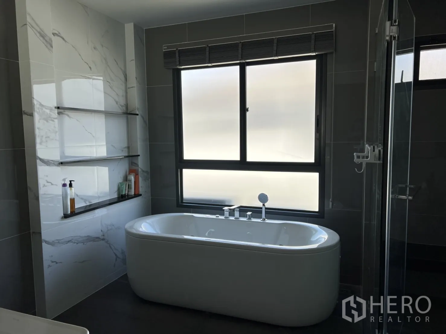 Detached House for rent or sale in Bang Kapi, Bangkok - Contemporary bathroom with freestanding soaking tub, glass shower and marble-effect wall tiles by a large frosted window.