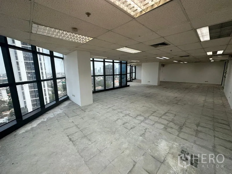 Office Space for rent - Bright unfurnished workspace with grid ceiling, fluorescent lighting, and full‑height windows facing high‑rise views.