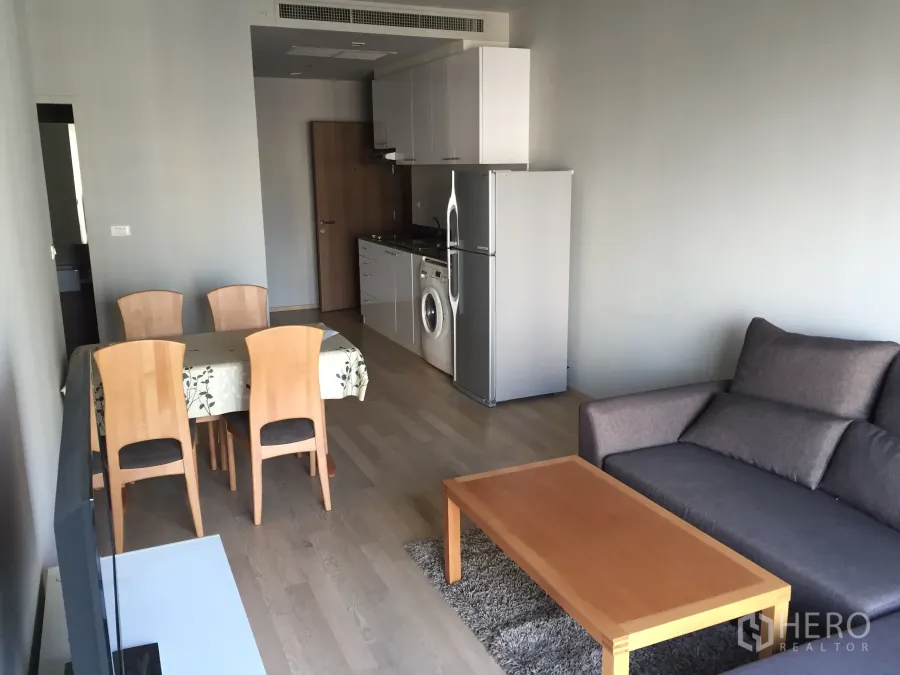Condominium for rent in Bangkok - Open-plan living and dining area with sofa, wooden coffee table, and kitchenette featuring a fridge and washing machine.