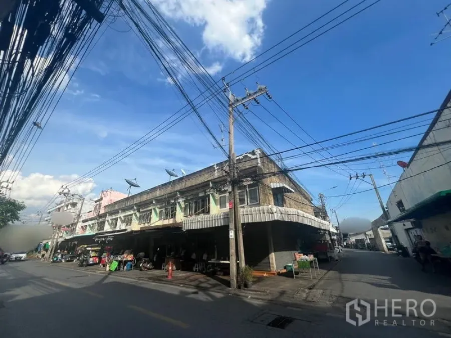 Land for rent or sale in Phra Khanong, Bangkok - Wide streetscape of a corner shophouse block with dual-road access and overhead wires in a neighborhood market area.