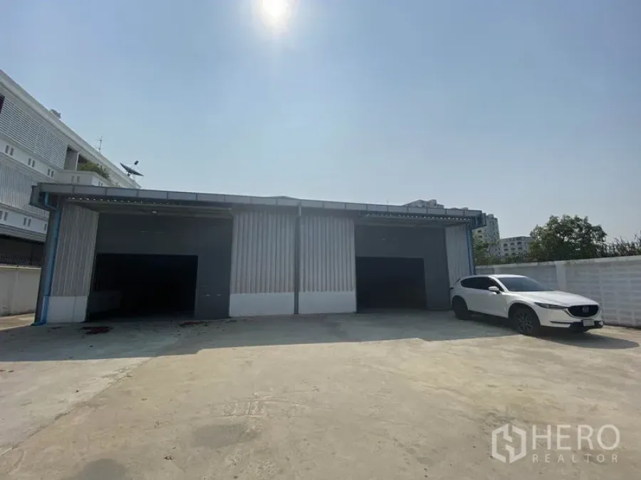 Warehouse for rent in Prawet, Bangkok - Metal-clad warehouse exterior with three roller shutters and a wide concrete yard, with a white SUV parked.