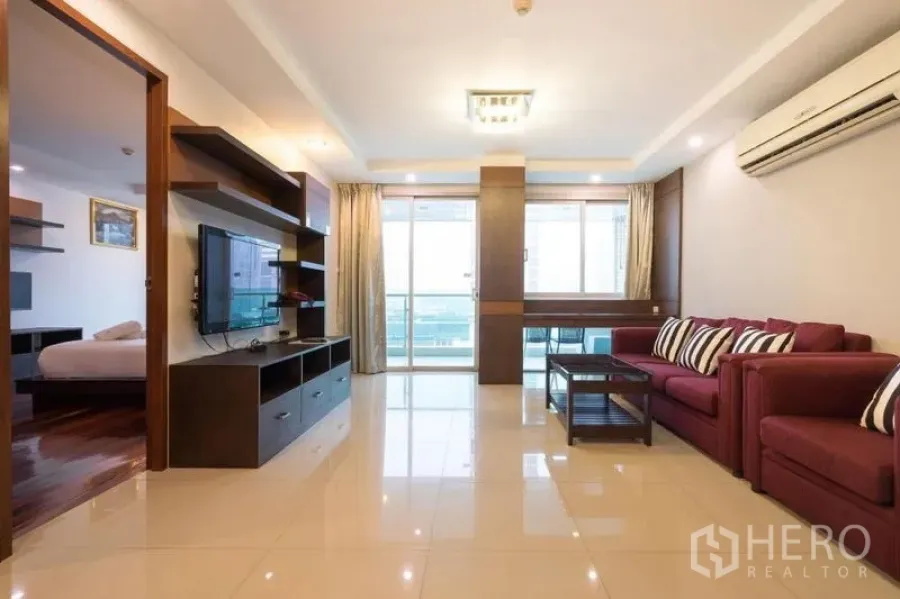 Condominium for rent in Watthana, Bangkok - Living area with maroon sofa set, TV console, shelving and access to a balcony.