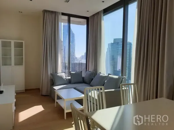 Condominium for rent in Pathum Wan, Bangkok - Corner living room with floor-to-ceiling windows, grey sofa and panoramic city views.