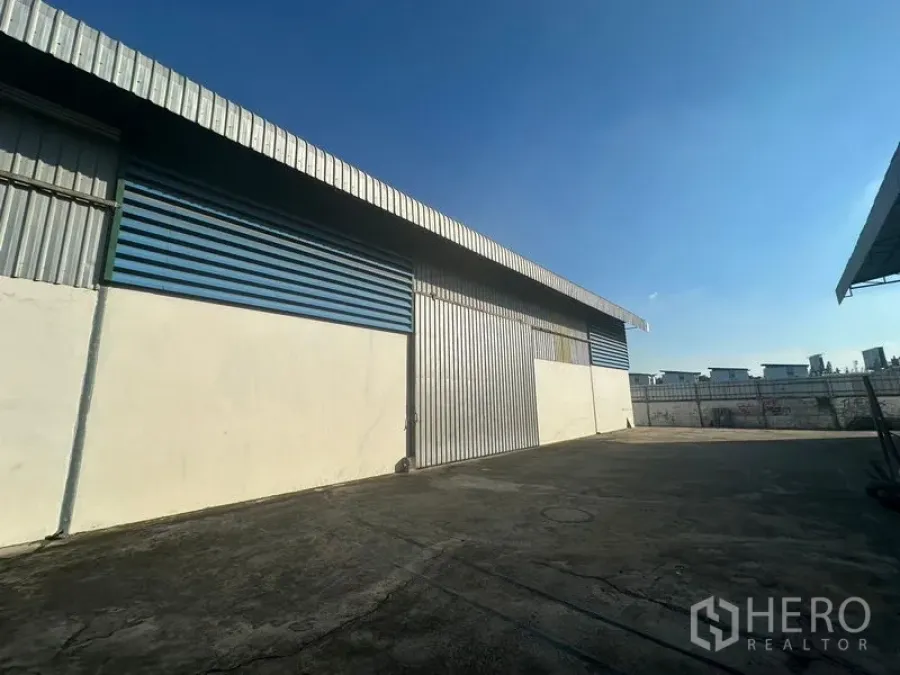 Warehouse for rent in Saphan Sung, Bangkok - Exterior of corrugated metal warehouse with large sliding door and wide concrete yard under blue sky.