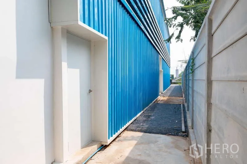 Warehouse for rent in Khlong Luang, Pathum Thani - Narrow side walkway along the warehouse with a white service door and boundary wall.