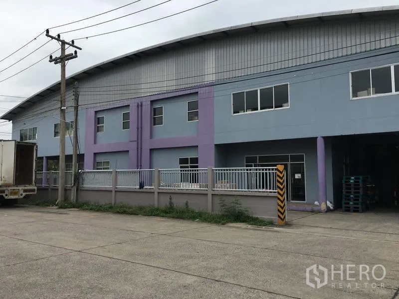 Factory for rent in Pathum Thani - Street-side view of a gated two-storey factory with pastel facade and ample parking apron.
