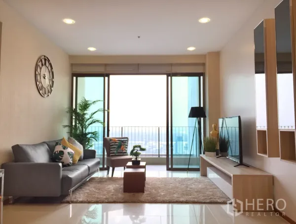 Condominium for rent or sale in Khlong Toei, Bangkok - Bright living room with grey sofa, rug and TV opening to a balcony with city view.