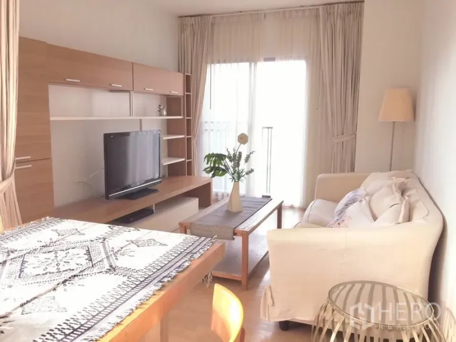 Condominium for rent in Bangkok - Bright living room with white sofa, wooden media wall, TV and sheer curtains opening to a balcony.