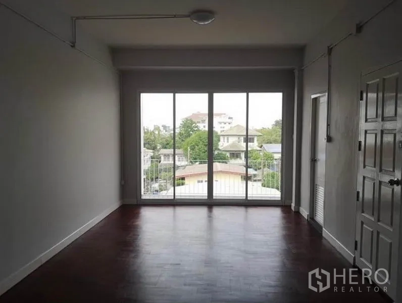 Townhouse for rent or sale in Din Daeng, Bangkok - Bright living room with floor-to-ceiling sliding glass doors opening to a balcony and city view, dark wood floors.