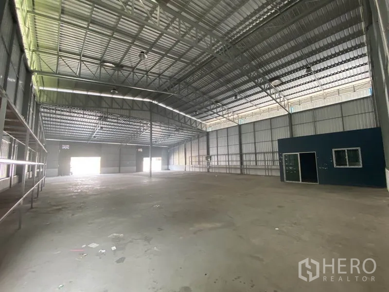 Warehouse for rent in Prawet, Bangkok - Spacious open-plan warehouse interior with high steel truss roof, skylight panels, and a small office cabin.