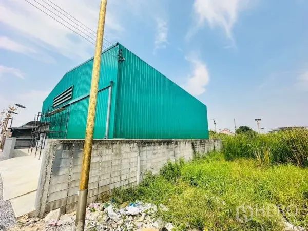 Warehouse for rent in Bang Phli, Samut Prakan - Green metal-clad warehouse exterior with ventilation louver and boundary wall beside a grassy area in Samut Prakan.