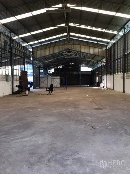 Warehouse for rent in Samut Prakan - Spacious clear-span warehouse floor with concrete slab under a metal truss roof allowing natural light.