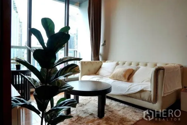 Condominium for rent in Khlong Toei, Bangkok - Bright living room with cream tufted sofa, round coffee table, indoor plant, and large window with city view.