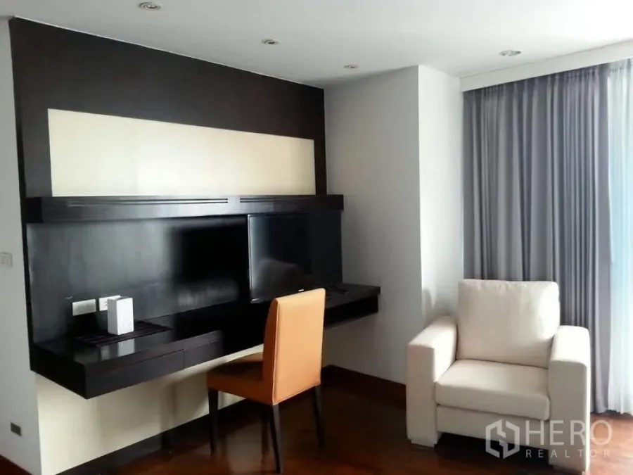 Condominium for rent or sale in Pathum Wan, Bangkok - Living area with floating TV wall, work desk, armchair, and floor‑to‑ceiling curtains.