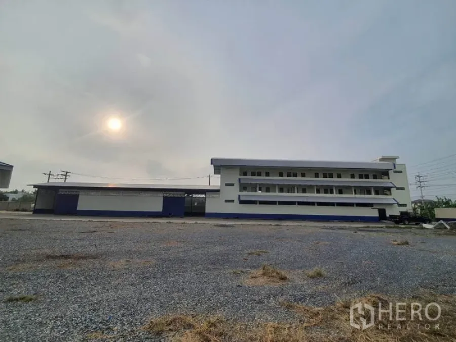 Factory for rent in Bang Bua Thong, Nonthaburi - Wide gravel yard facing a multi-storey factory and office block under a hazy sky.