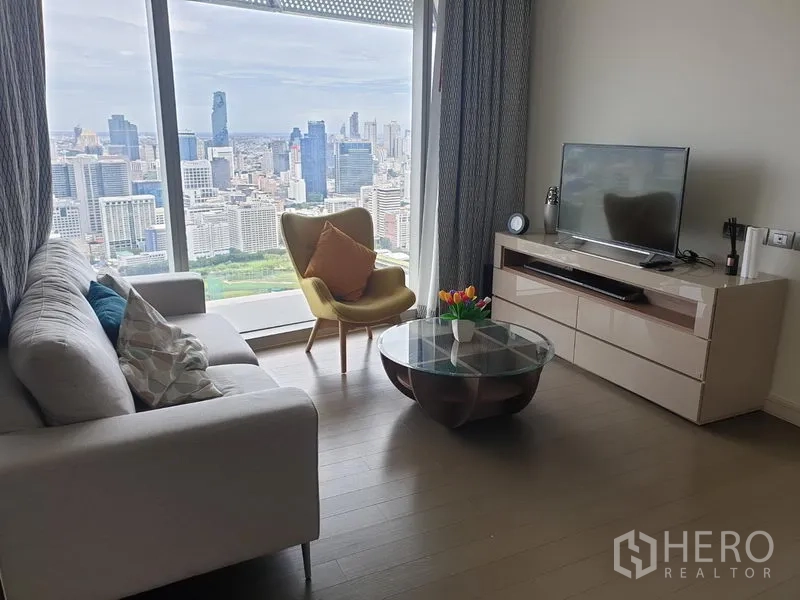 Condominium for rent in Pathum Wan, Bangkok - Living room with TV console and glass coffee table facing floor-to-ceiling windows and skyline views.