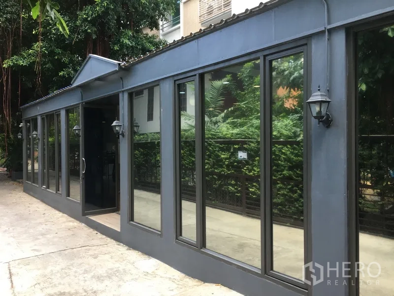 Retail Space for rent in Watthana, Bangkok - Street-facing single-storey retail unit with dark metal facade and large glass windows beside lush greenery in the Thonglor area of Watthana, Bangkok.